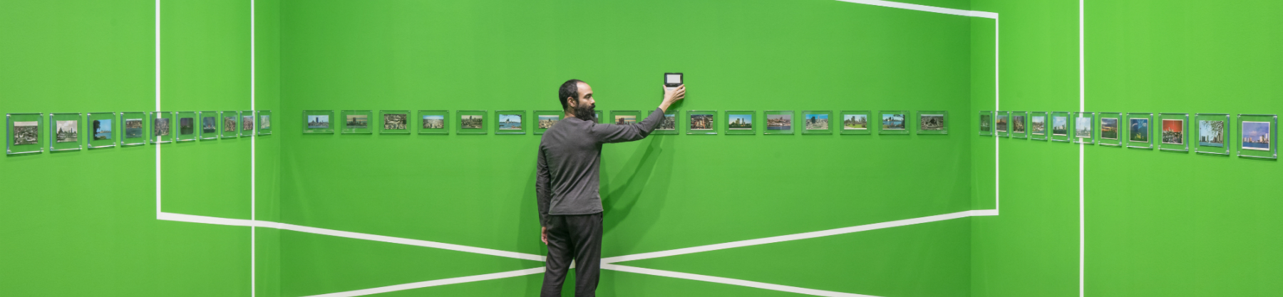 A man stands in the middle of a gallery of photographs. The walls are bright green and the photos are hung in a horizontal line around the room.