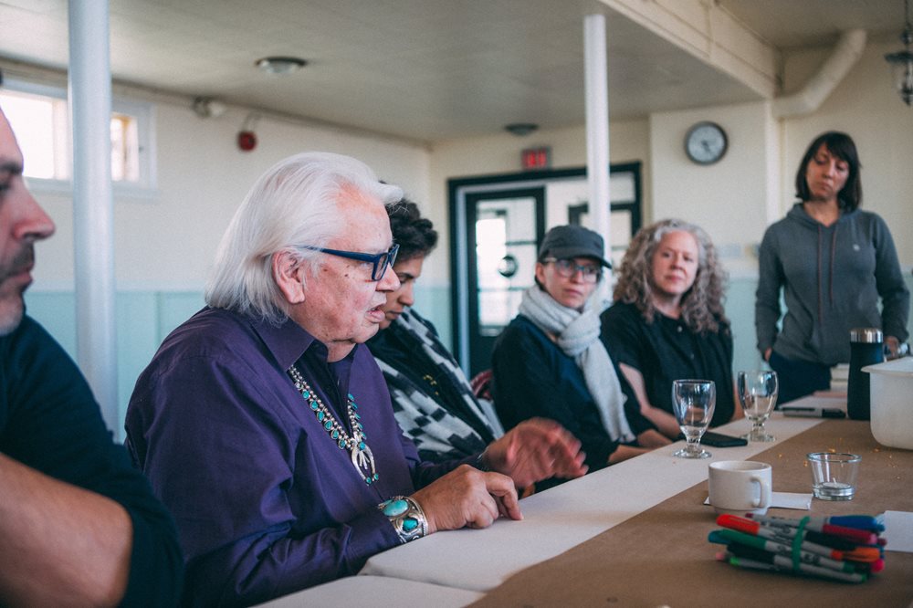 A group of people sit along a table listening to Elder Duke Redbird speak