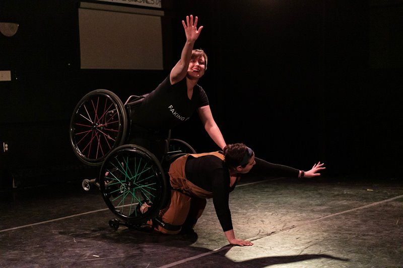 A colour photo of two dancers strapped into wheelchairs. Jen, on hands and knees on Shay’s back. Each dancer gazes at their extended arm, smiling