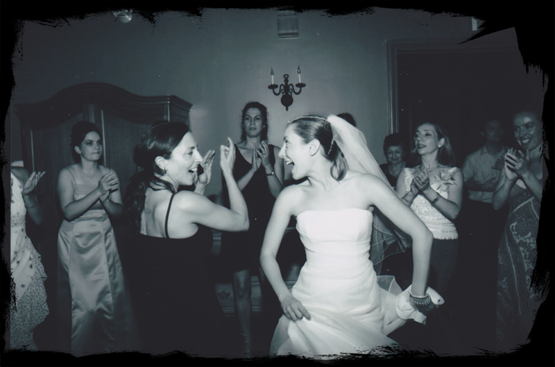 Black and white photo of two women; a bride, dressed in white with a veil, another in a black party dress, performing a Spanish folk dance, Sevillanas, surrounded by joyful wedding guests