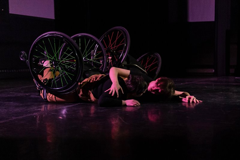 Colour photo of two dancers strapped to wheelchairs, laying face down on the floor. Jen’s arm is wrapped around Shay’s shoulder as they look intensely at each other.